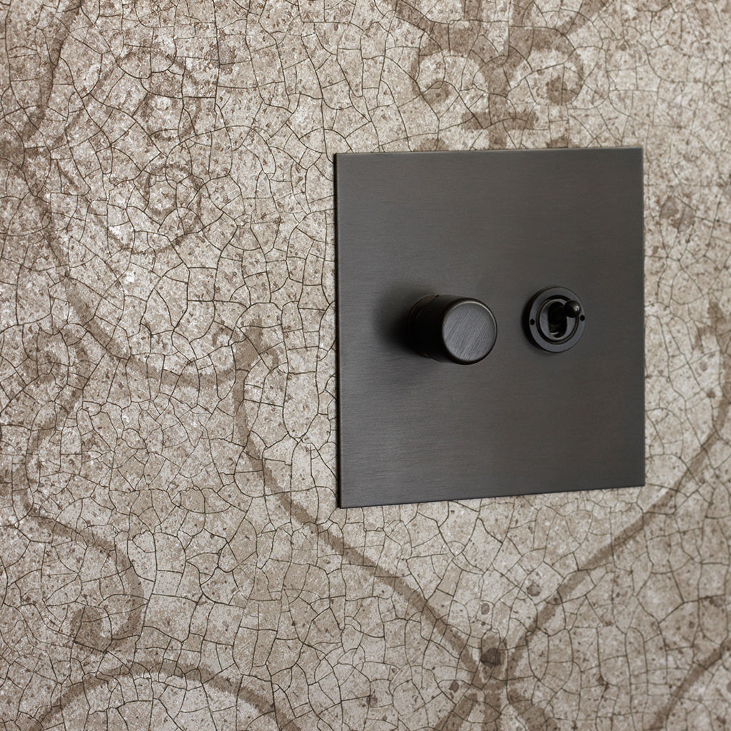 A bronze Forbes & Lomax switch plate on patterned wallpaper.