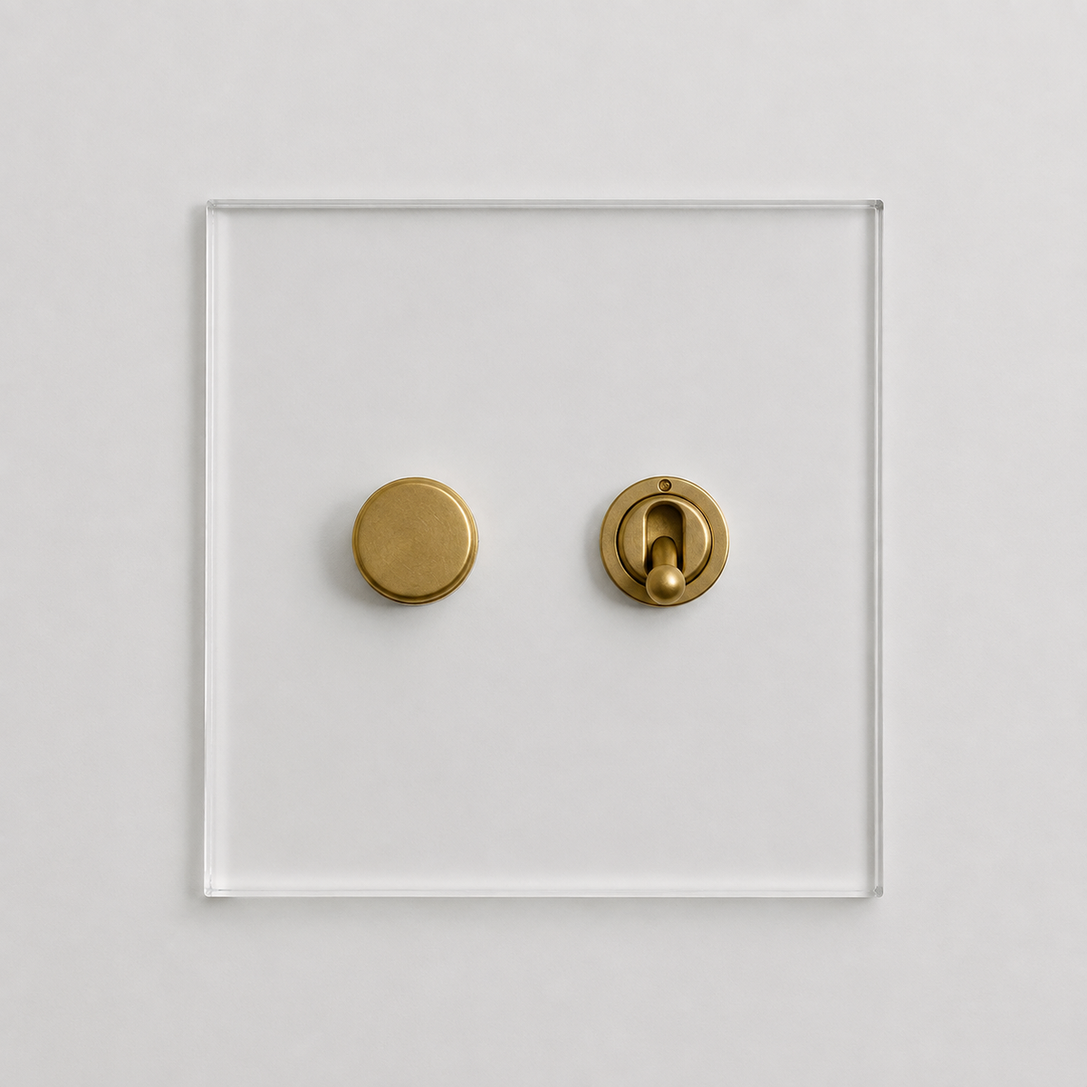 A modern two-button switch plate featuring aged brass components, set against a clear square acrylic panel on a light gray background.