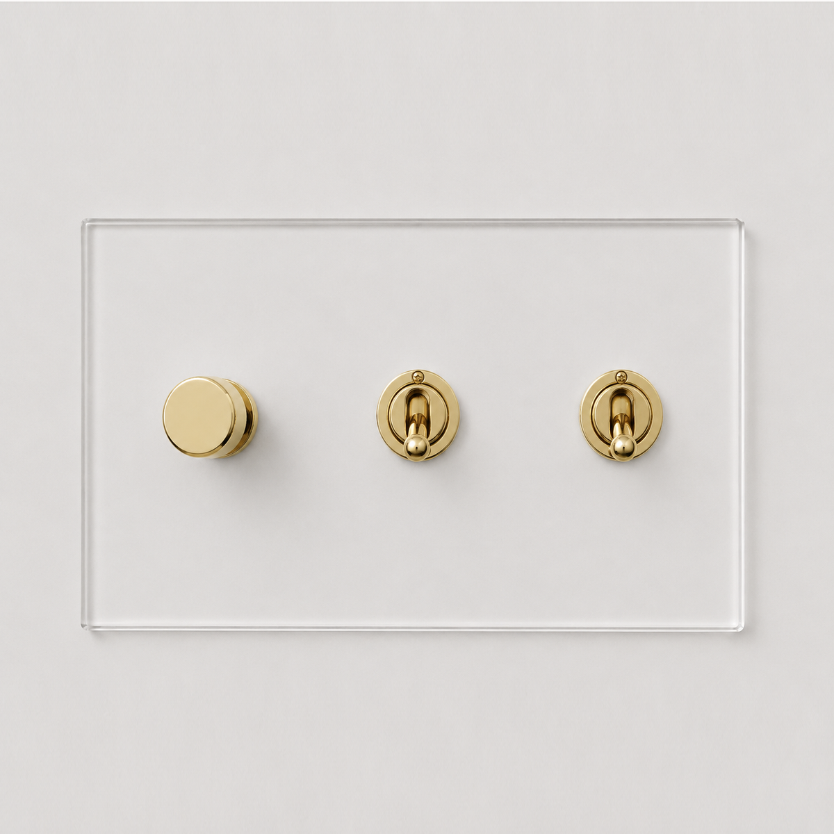 Forbes & Lomax invisible acrylic plate with one brass rotary dimmer and two brass toggle switches on a white wall.