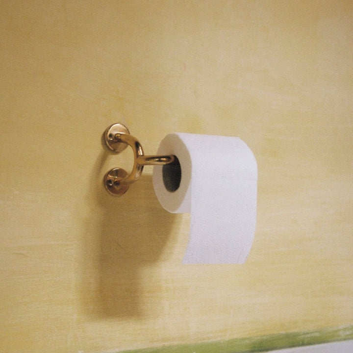 Fauna Toilet Roll Holder in polished brass installed on a textured butter yellow wall with a roll of toilet paper mounted.