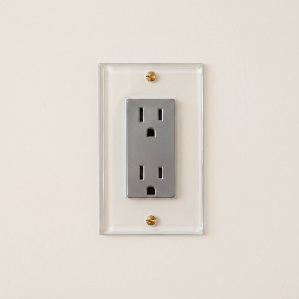 Forbes & Lomax Invisible outlet with a silver receptacle on a transparent acrylic plate mounted on a beige wall.