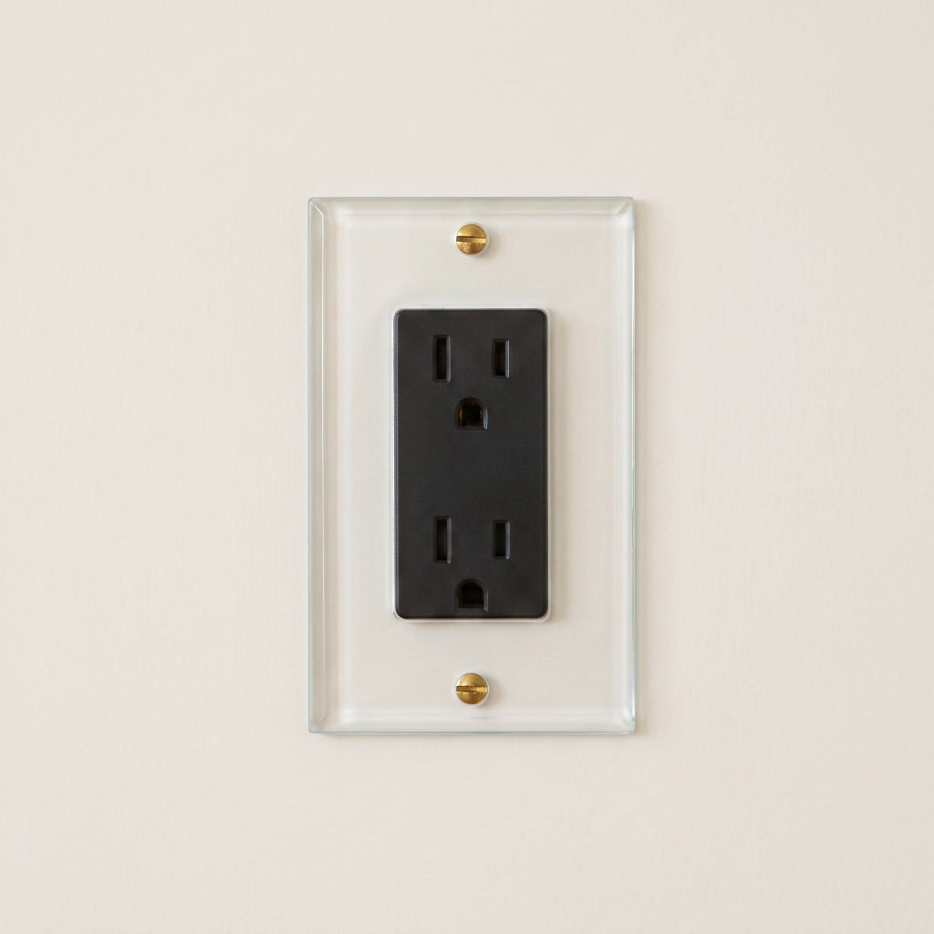 Forbes & Lomax Invisible outlet with a black receptacle on a clear acrylic plate and brass screws on a beige wall.
