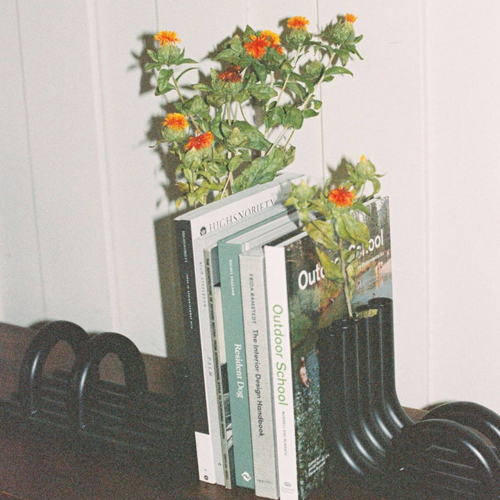 Black TUBE Bookends photographed on a wood table in a white room with a collection of books in between and flowers inside the tubes