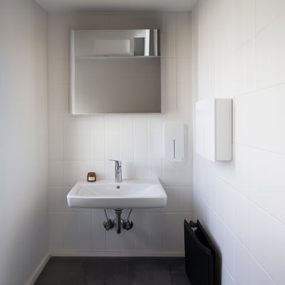 Loki Hand Dryer and Soap Dispenser in white installed in a commercial bathroom against white tiled walls.
