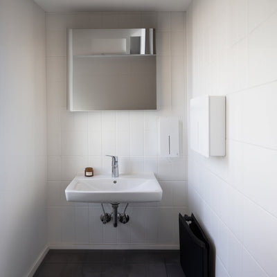 Loki Soap Dispenser and Hand Dryer in white installed in a commercial bathroom against white tiled walls.