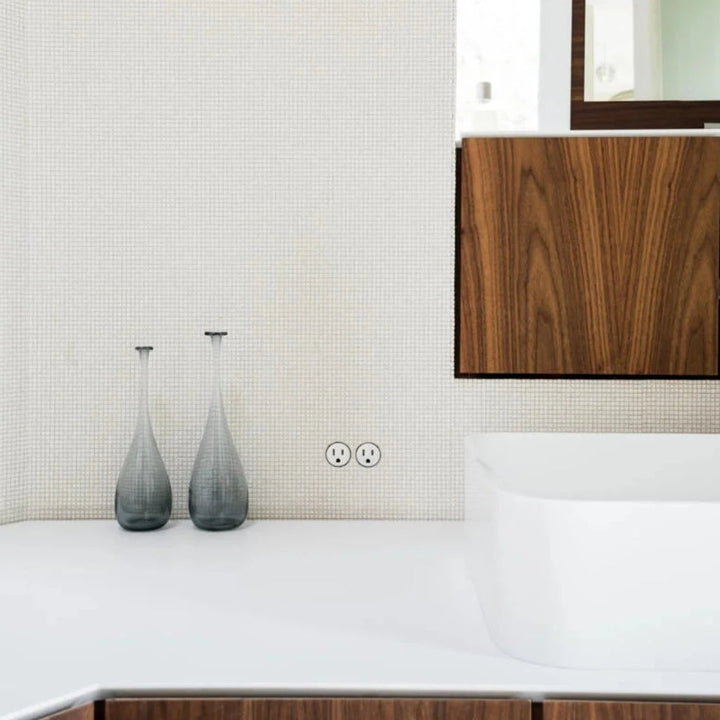 An electrical outlet mounted flush into a mosaic tile surface in a bathroom. 
