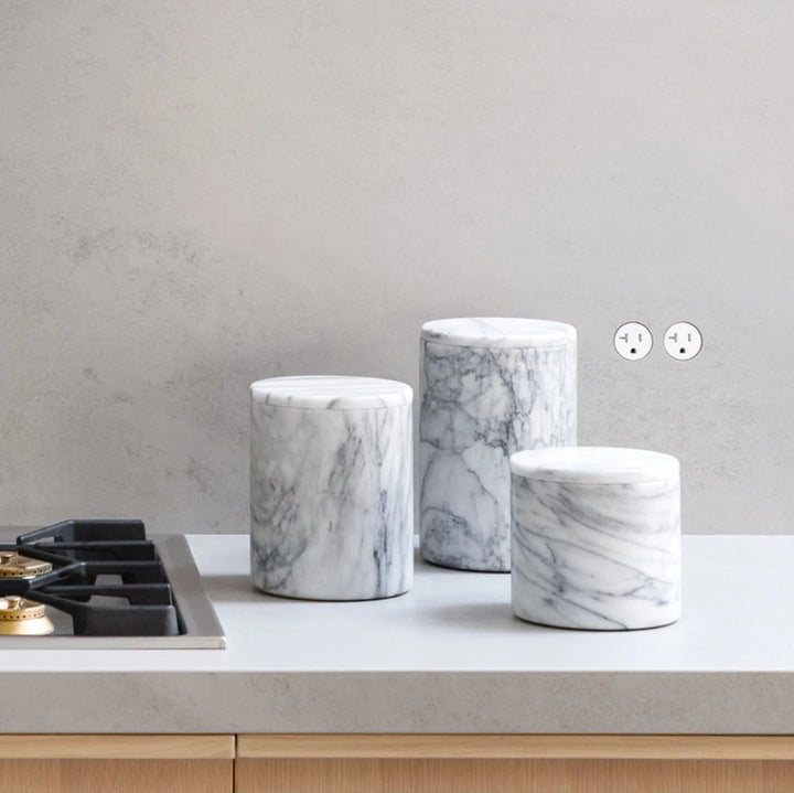 Marble-patterned containers on a kitchen counter with gas cooktop and flush Bocci22 outlets in a plaster backsplash wall. 