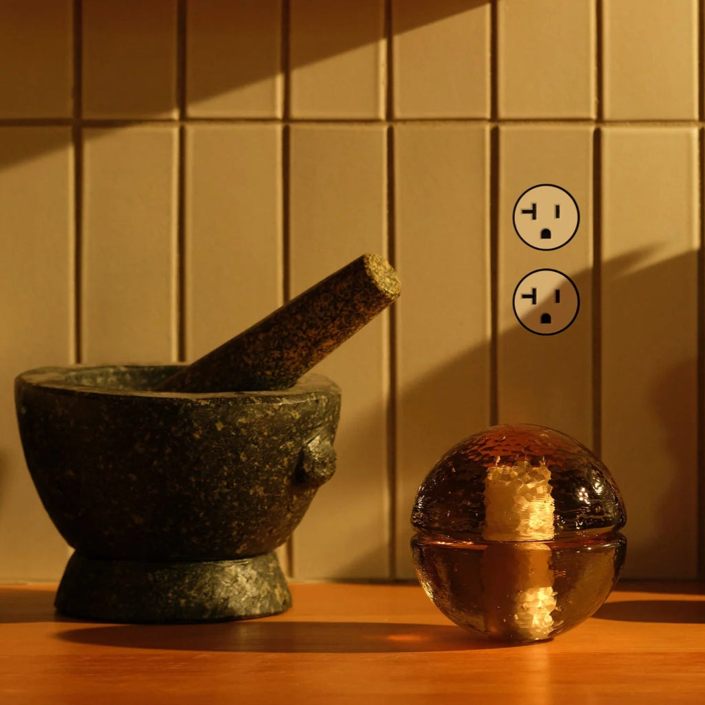 A flush wall socket cover plate is shown in a partially illuminated setting, with a mortar and pestle on the left and a round table lamp on the counter in  kitchen. 