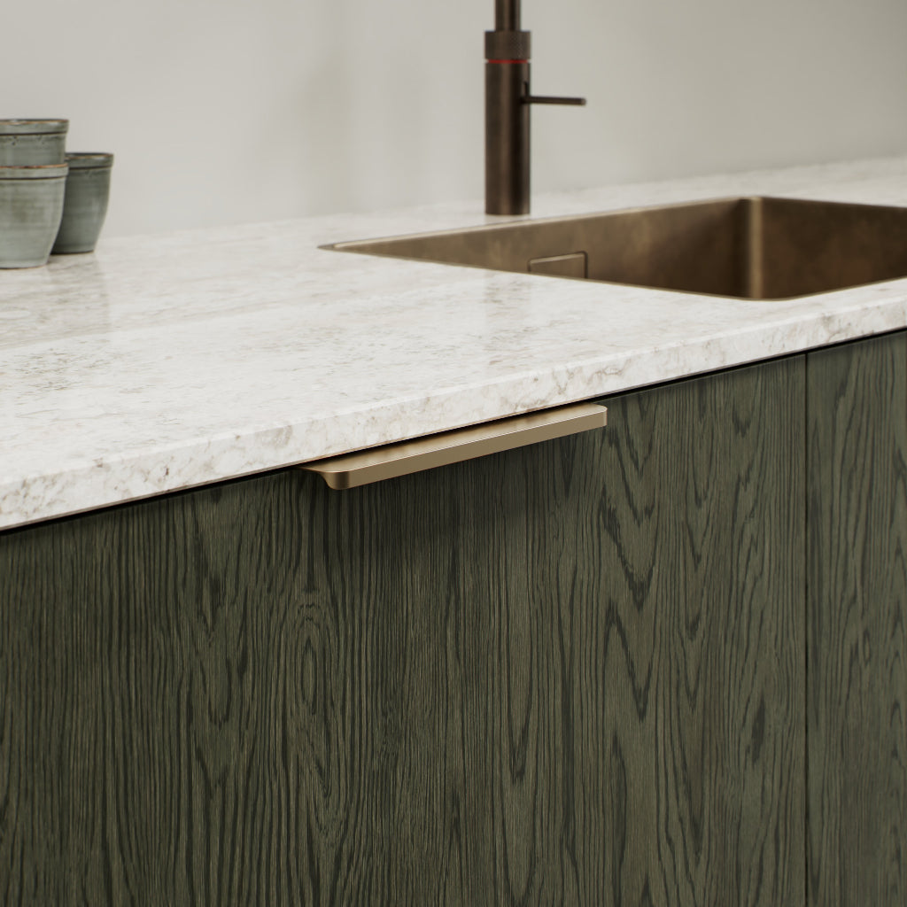 Matte brass Furnipart Comet edge pull, 160 mm, on a dark green oak cabinet door under a marble countertop.