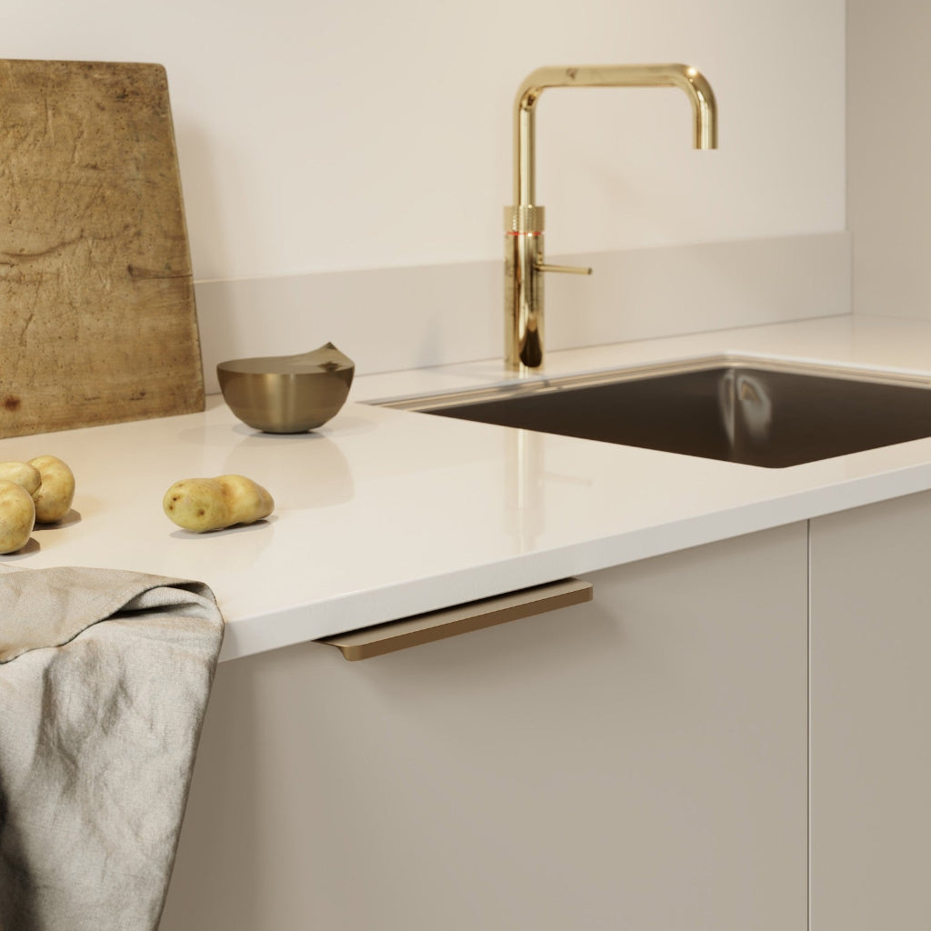 Matte brass Furnipart Comet edge pull, 160 mm, mounted at the top of a beige cabinet door under a white countertop.