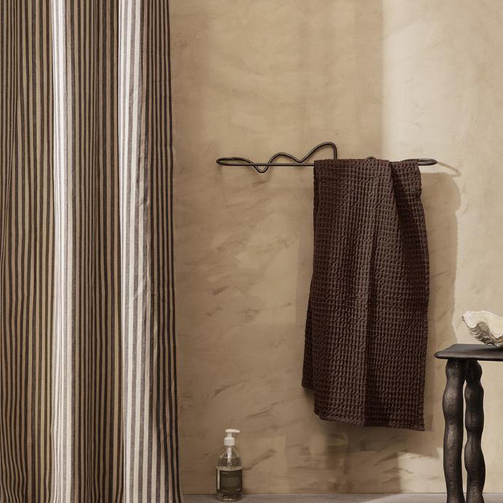 A Ferm Living Curvature Towel Bar hanging on a hook next to a shower curtain.
