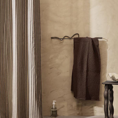 A Ferm Living Curvature Towel Bar hanging on a hook next to a shower curtain.