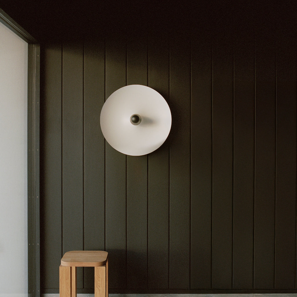 Round wall light fixture on a dark paneled wall with a wooden stool in the foreground.