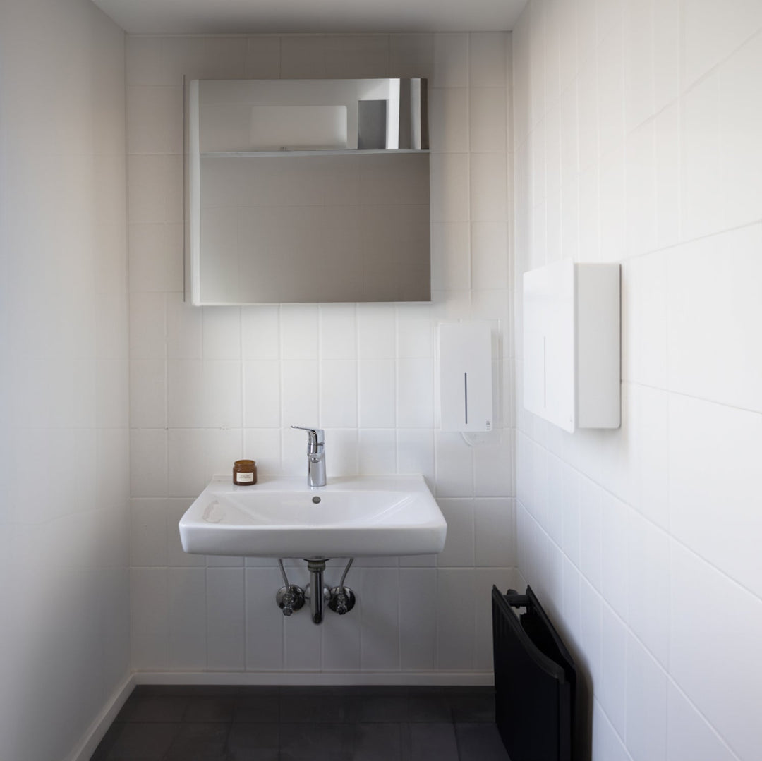 Loki Hand Dryer and Soap Dispenser in white installed in a commercial bathroom against white tiled walls.