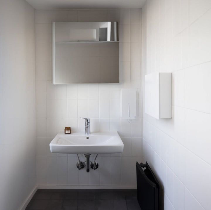 Loki Soap Dispenser and Hand Dryer in white installed in a commercial bathroom against white tiled walls.