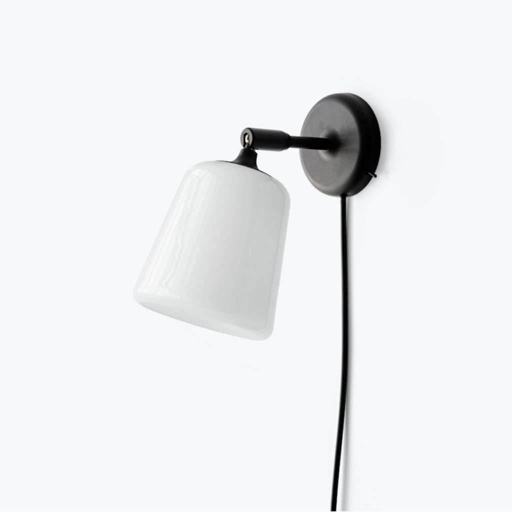 Minimalist wall lamp with a matte black frame and a glossy white glass shade on a pivoting head against a white wall.
