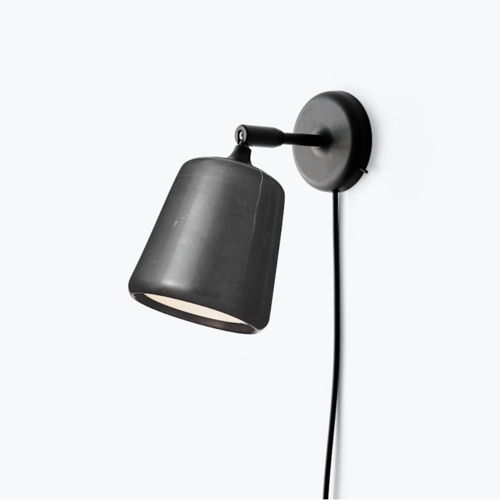 Minimalist black wall lamp with a pivoting dark stone shade and cord, mounted on a plain white background.