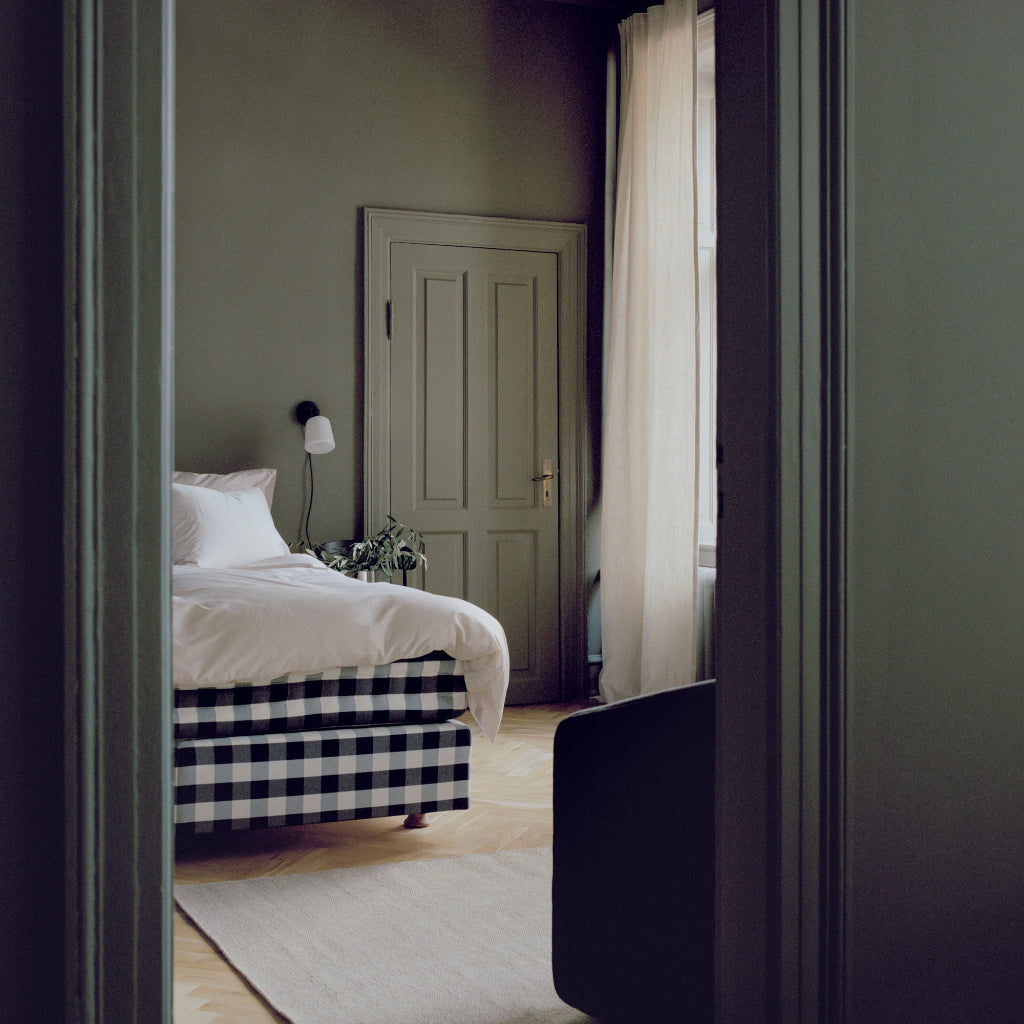 Minimalist white wall lamp with a black frame mounted in a cozy bedroom with sage green walls and a checkered bed base.