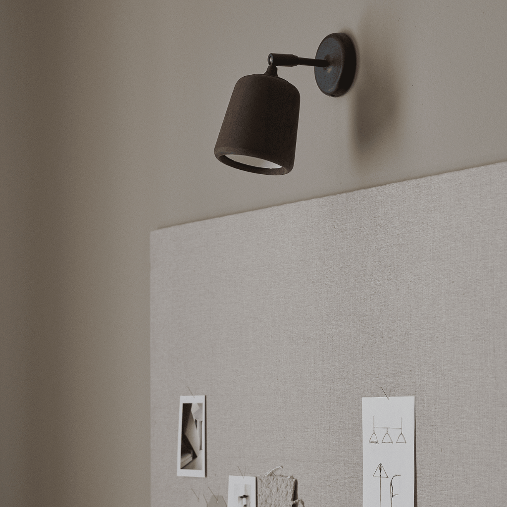 Minimalist black wall lamp with a pivoting head mounted above a beige fabric pinboard in a neutral room.