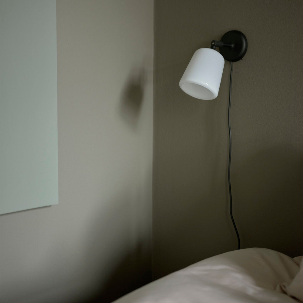 Minimalist wall lamp with a white glass shade and black frame mounted on a dark grey bedroom wall.