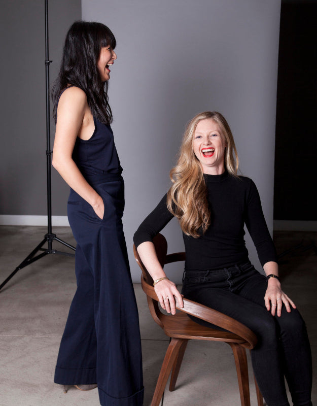 Megan Cassidy and Jane Son posing in a studio setting. One woman stands on the left wearing a dark navy sleeveless maxi dress, and the other sits on a wooden chair on the right wearing a black long-sleeve top and black pants.