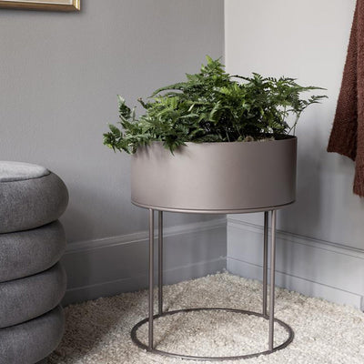 A Ferm Living Plant Box Round on a stand in a corner of a room.