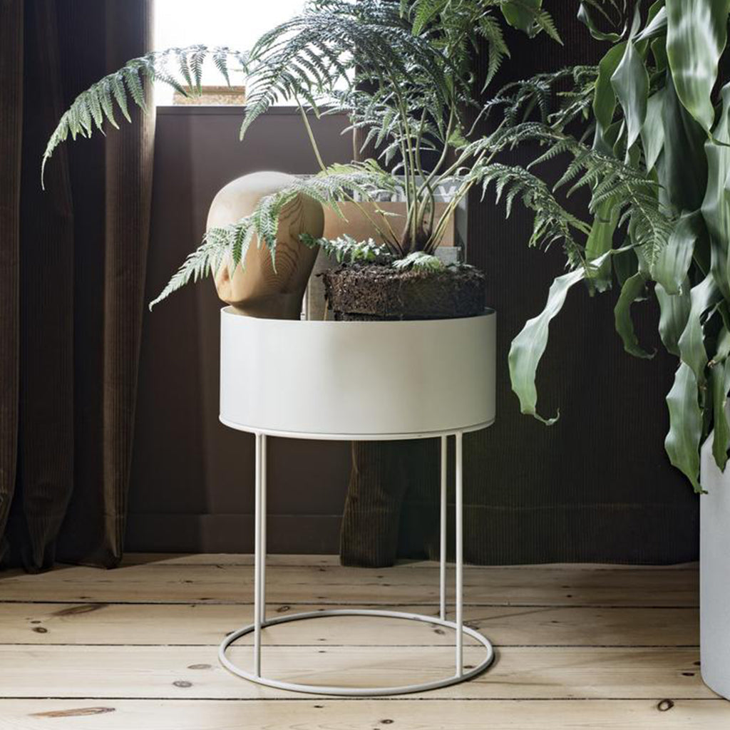 A Ferm Living Plant Box Round sits on a stand next to a window.