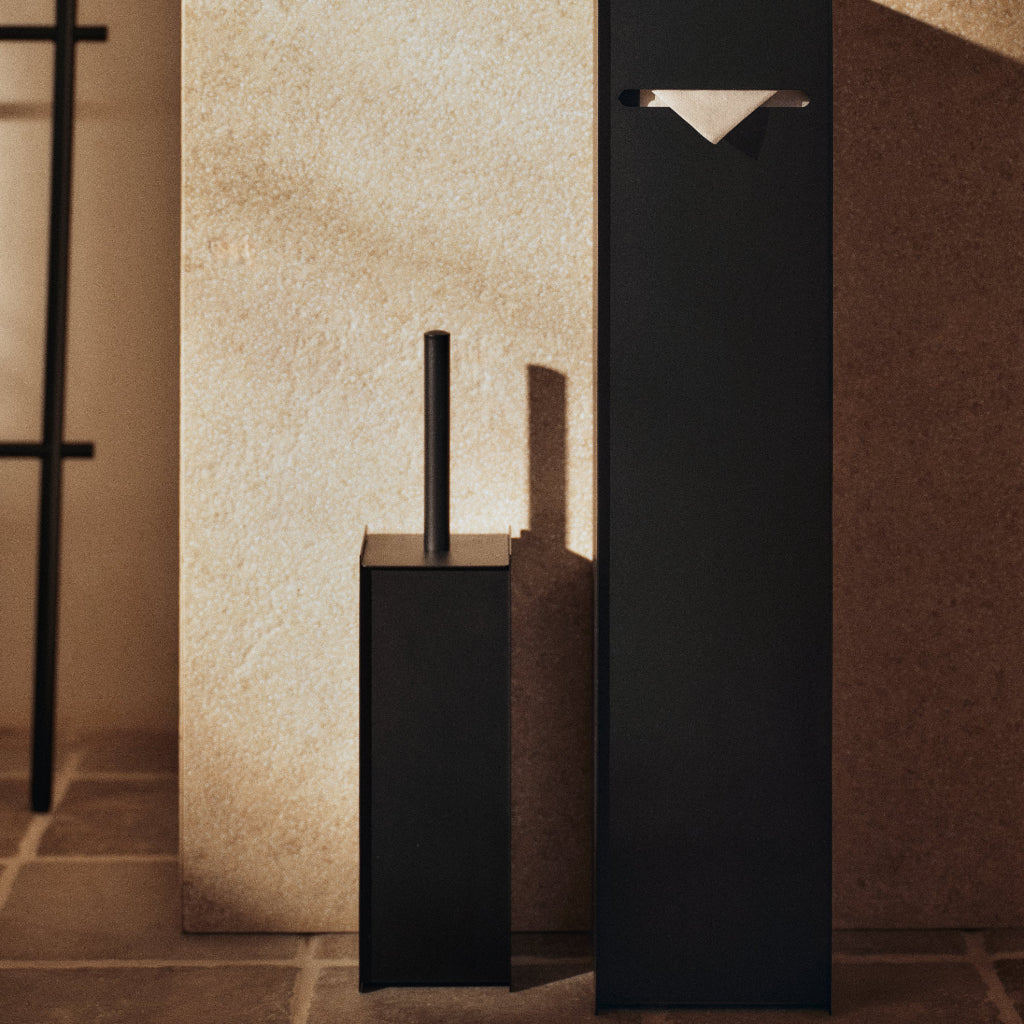 Black Sama toilet brush in a sleek, cylindrical stainless steel holder against a textured stone wall.