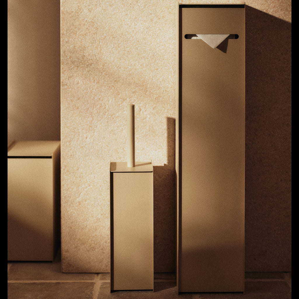 Modern brushed stainless steel Sama toilet paper dispenser in cashmere finish against a textured stone wall.