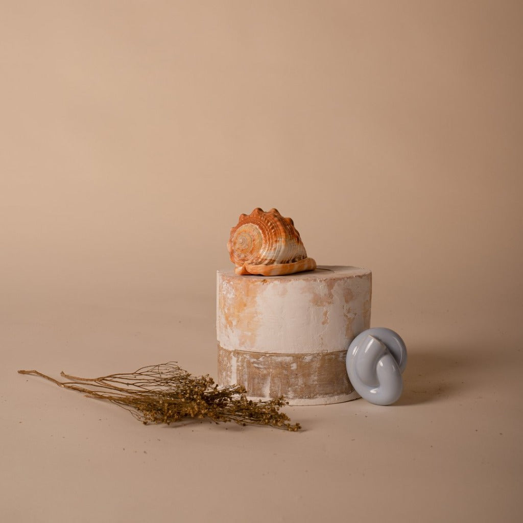 A blue ceramic wall hook or pull softly shaped like a cord tied into a knot, resting against an organic shaped prop against a neutral sand coloured background.